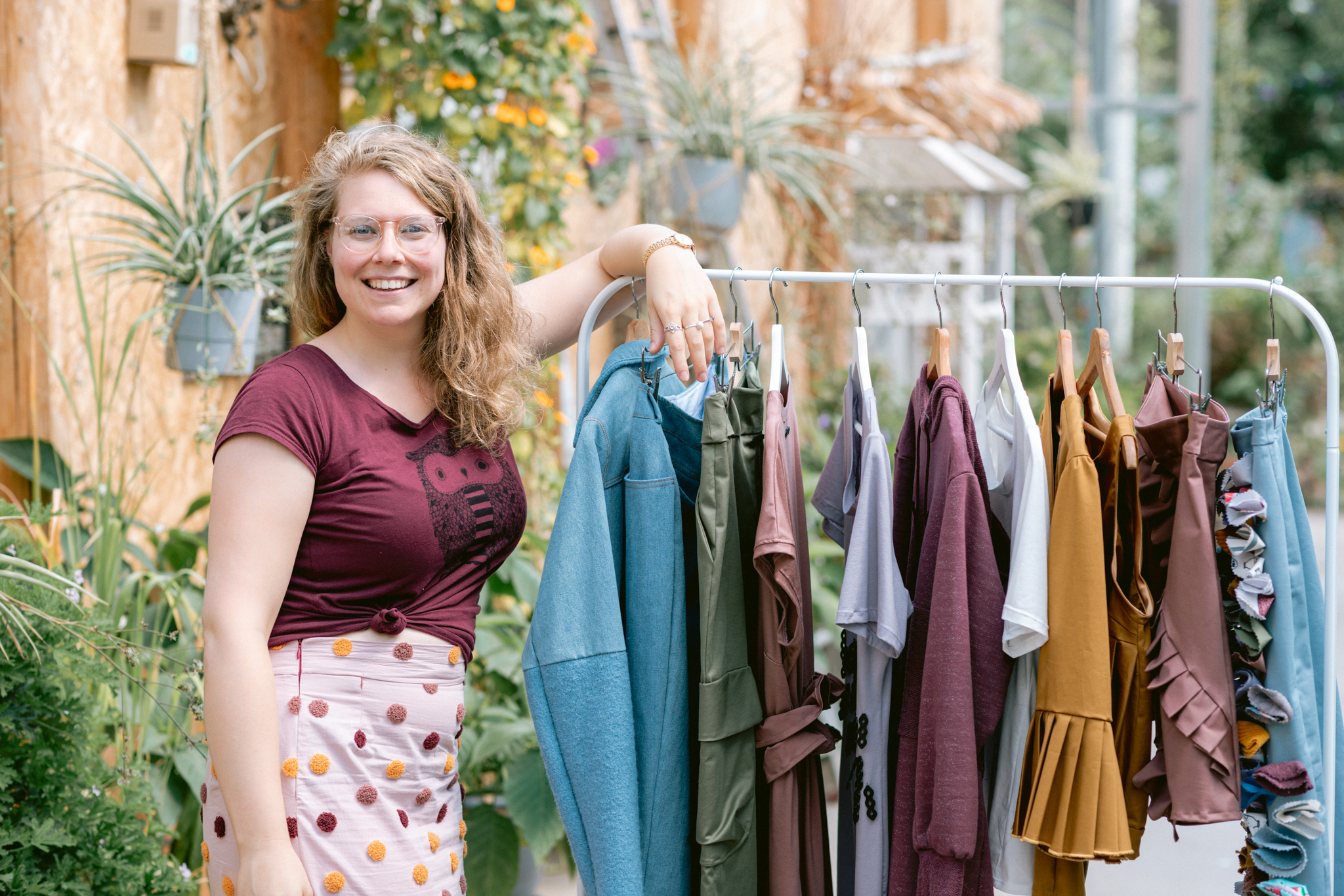 Rachel staat naast een kledingrekje vol SeeFeel kleding. Ze draagt een bordeauxrood T-shirt en een roze rok met stippen