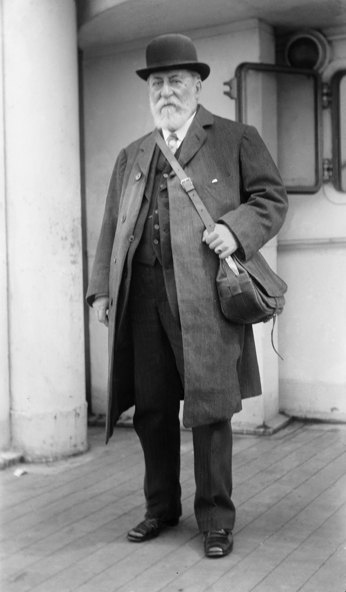 De componist Camille Saint-Saëns staat buiten in reiskleding met een bolhoed en reistas.