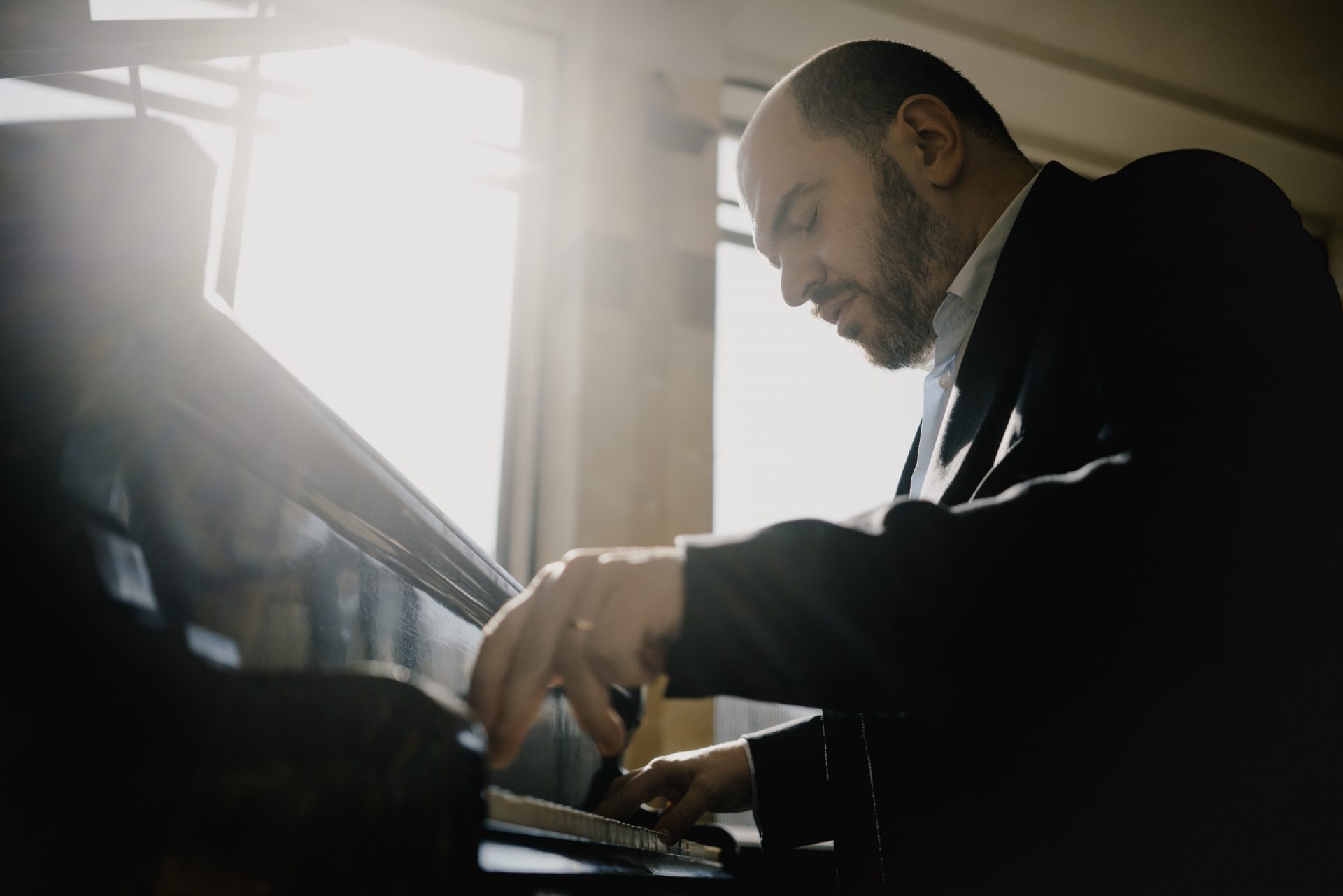 Pianist Kirill Gerstein speelt geconcentreerd piano voor een raam waar de zon doorheen schijnt.